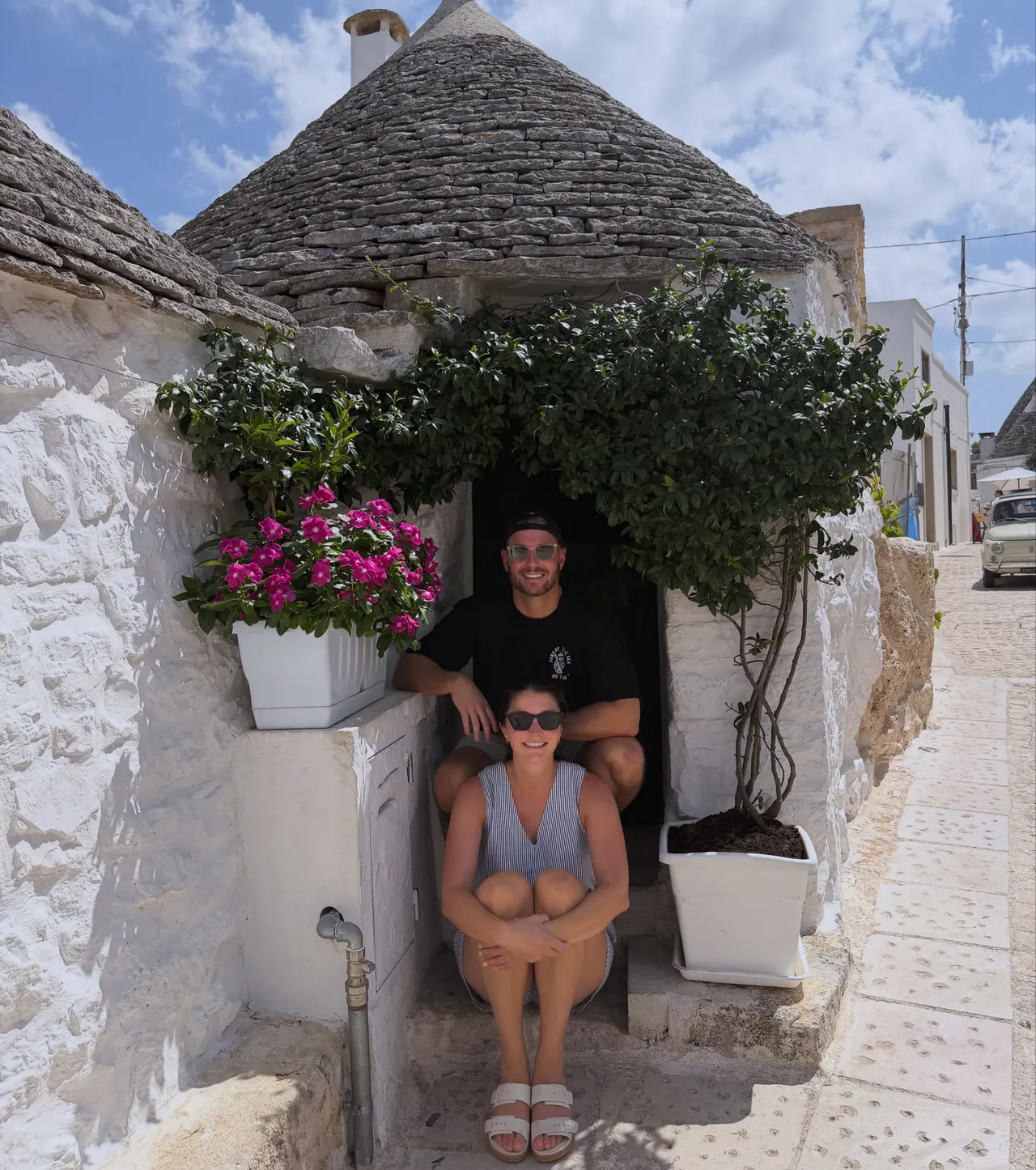 Drew and Valerie seated in the doorway of a trullo framed by pink flowers